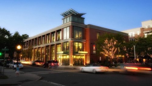 A modern building with large windows, illuminated at dusk, surrounded by trees and passing cars.