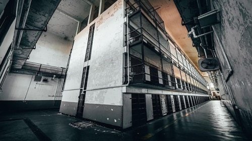 Abandoned prison interior with empty cell blocks and peeling walls, illuminated by overhead lights.