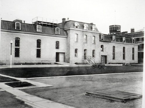 Historic black-and-white photo of a stone building with a sloped roof and large windows, surrounded by a grassy area.
