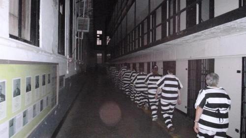 A dimly lit prison corridor with inmates in striped uniforms walking away from the camera.