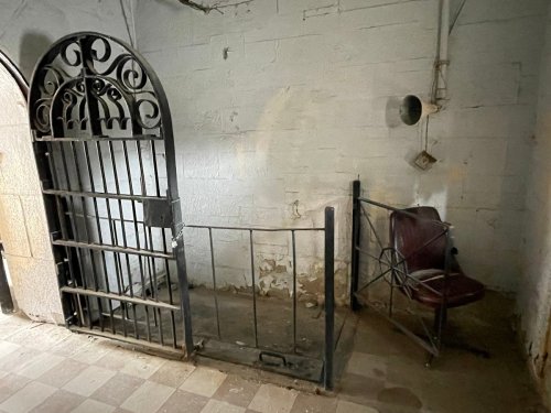A dimly lit room featuring a barred gate and a solitary metal chair against a bare wall.