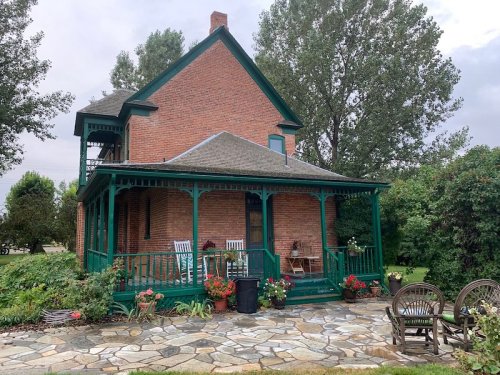 A charming brick house with a green porch, surrounded by flowers and a stone patio with wooden chairs.