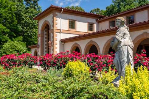 A stone statue stands in a garden filled with colorful flowers, in front of a beautiful building with arched windows.
