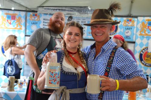 A man and woman in traditional attire hold beer mugs at a festival, with a smiling person in the background.
