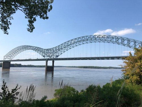 A large, arched bridge spans a river, surrounded by greenery and a clear blue sky.
