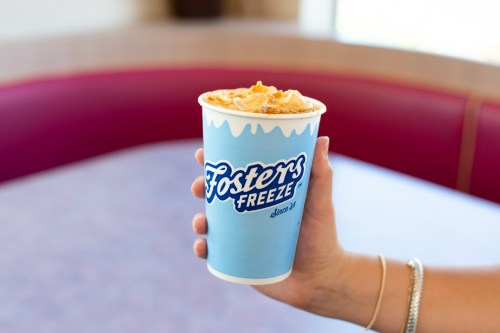 A hand holding a blue cup from Foster's Freeze, topped with whipped cream and caramel, in a casual dining setting.