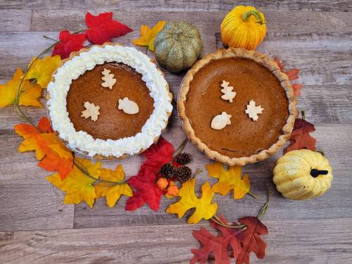 Two pumpkin pies decorated with fall-themed cutouts, surrounded by colorful autumn leaves and small pumpkins.