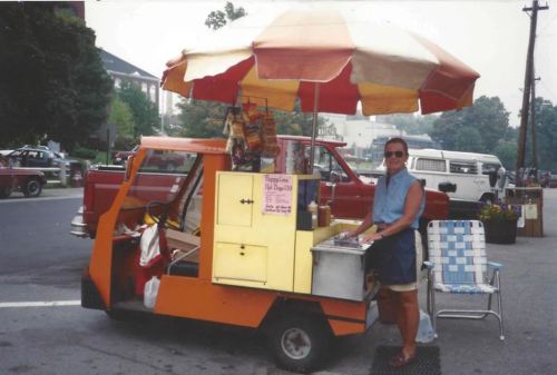 A vendor stands by a colorful food cart with an umbrella, serving snacks on a sunny street.