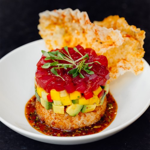 A colorful sushi stack with tuna, mango, and avocado, served on a plate with crispy rice and sauce.