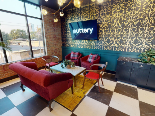 Cozy lounge area with a red sofa, plaid chairs, a coffee table, and decorative wallpaper, featuring a "puttery" sign.