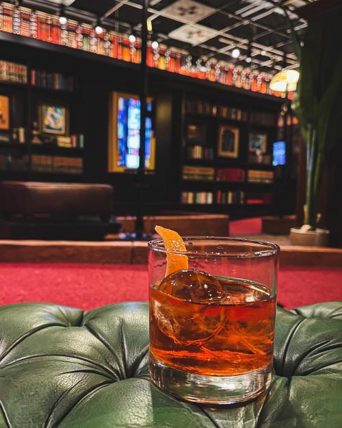 A close-up of a cocktail glass with an orange twist, set on a green leather surface, with a library backdrop.