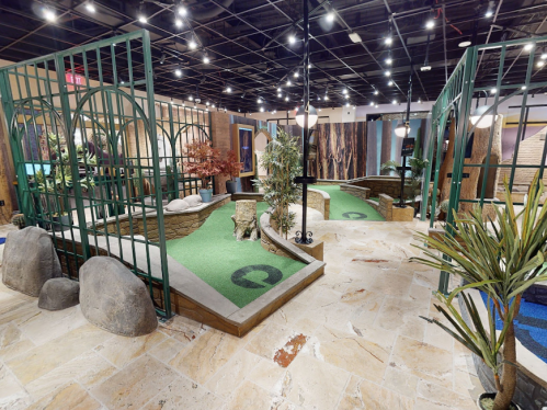 A vibrant indoor mini-golf course featuring greenery, rocks, and decorative plants under bright lights.