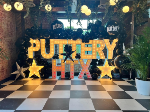 Brightly lit "PUTTERY HTX" sign with black balloons and decorative stars in a stylish indoor setting.