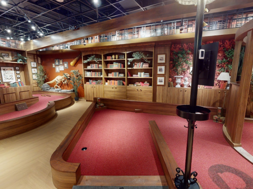 A cozy, warmly lit room with wooden shelves, red carpet, and decorative plants, resembling a library or reading area.