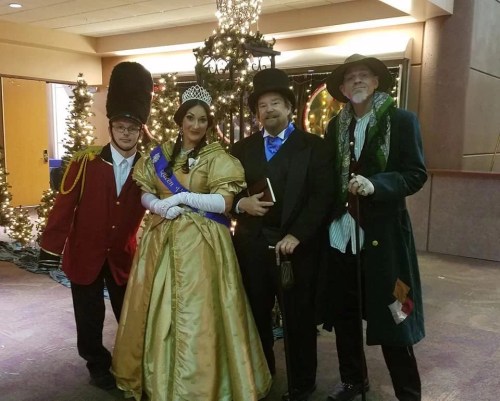 Four people in festive attire pose together in front of a decorated backdrop with Christmas lights and trees.