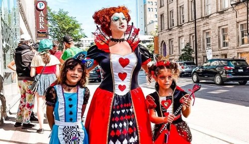 A woman dressed as the Queen of Hearts stands with two girls in costume, smiling on a city street during a festival.