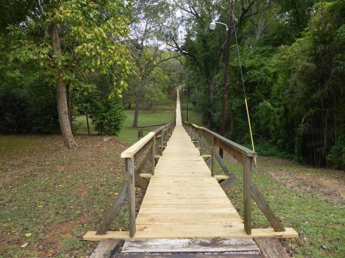 A wooden bridge leads through a green landscape, surrounded by trees and grass, towards a distant path.