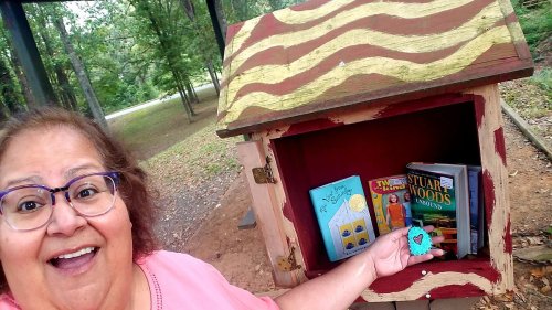A smiling woman holds a small heart-shaped object in front of a colorful little free library filled with books.