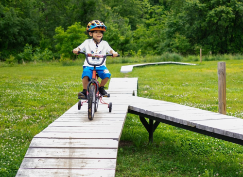 A young boy rides a bike on a wooden path through a grassy area, surrounded by trees.