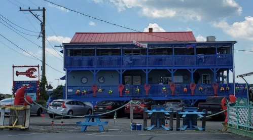 A blue seafood restaurant with a red roof, decorated with buoys, and surrounded by parked cars and outdoor seating.