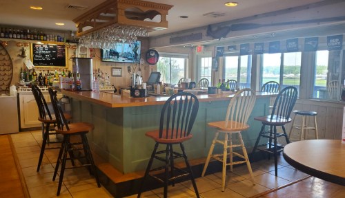 A cozy bar with a wooden counter, high stools, and a view of the outdoors, featuring shelves of drinks and a TV.