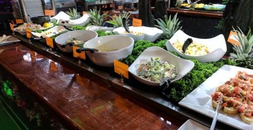 A buffet table with various salads and dishes, surrounded by greenery and labeled with orange signs.