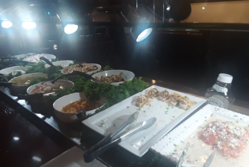 A buffet display with various dishes in bowls, garnished with lettuce, and serving utensils under bright lights.