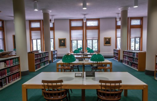 A quiet library interior with green lamps, wooden tables, and bookshelves, featuring large windows and portraits on the walls.