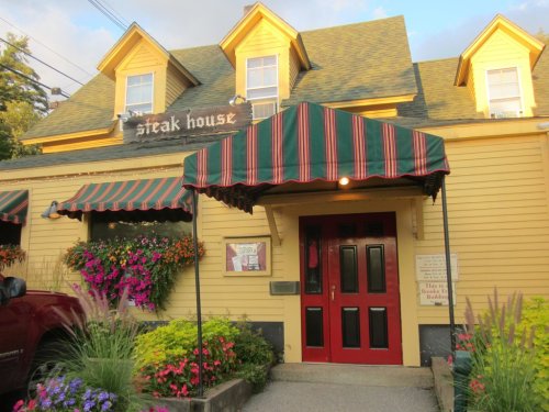 A yellow steakhouse with a red door and striped awning, surrounded by colorful flowers and greenery.