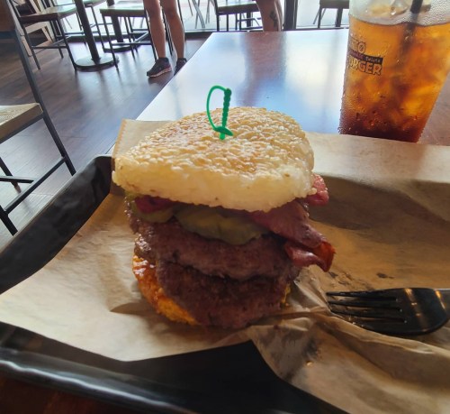 A burger with two patties, bacon, pickles, and a rice cake bun, served on a tray with a drink in the background.