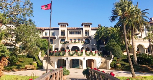 A grand mansion with palm trees, a flag, and festive decorations, surrounded by lush greenery and a wooden bridge.