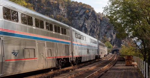 A silver train travels along a track near rocky cliffs and a tunnel, surrounded by trees in a scenic landscape.