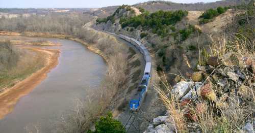 A train travels along a winding track beside a river, surrounded by hills and sparse trees in a rural landscape.