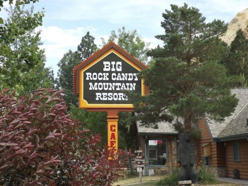Sign for Big Rock Candy Mountain Resort surrounded by trees and shrubs, with a rustic building in the background.