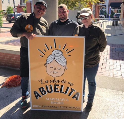 Three people stand beside a sign for "La Casa de Mi Abuelita," promoting traditional Mexican food.