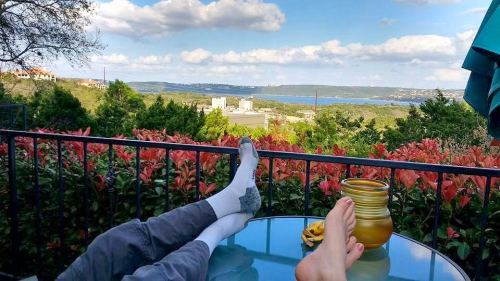 A scenic view of a lake and hills, with two pairs of feet resting on a table surrounded by colorful plants.