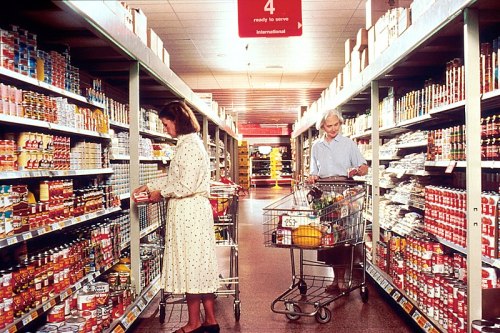 Two shoppers in a grocery store aisle, one selecting items from a shelf while the other pushes a cart.