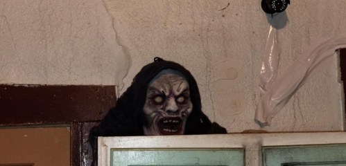 A creepy, masked figure with glowing eyes peeks over a door, set against a textured wall.