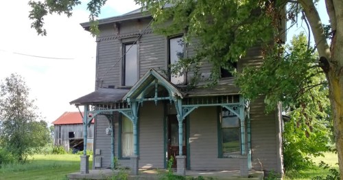 An old, two-story house with a porch, surrounded by trees and open land, showing signs of wear and age.