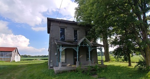A weathered two-story house with a porch, surrounded by greenery and fields under a partly cloudy sky.