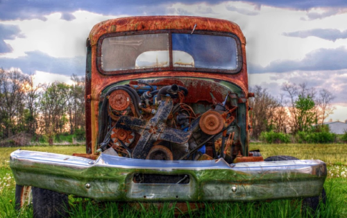 Rusty vintage truck with an exposed engine, set against a cloudy sky and green field.