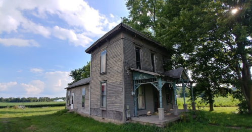 A weathered two-story house with a porch, surrounded by greenery and under a partly cloudy sky.