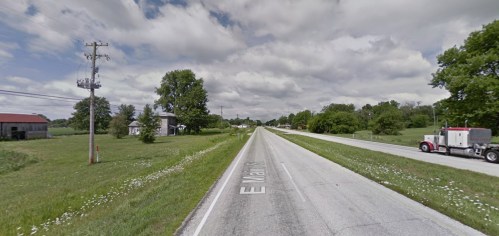 A rural road lined with green fields and trees under a cloudy sky, with a truck on the right side.