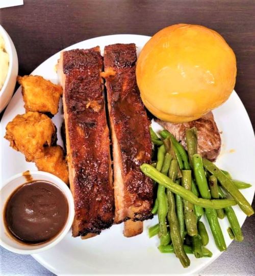 A plate of food featuring ribs, green beans, a roll, fried chicken, and a side of sauce.