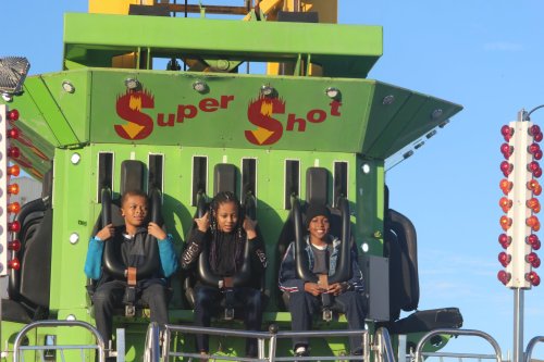 Three children sit in harnesses on a green amusement park ride called "Super Shot," ready for an exciting drop.
