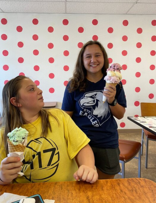 Two girls in a colorful ice cream shop, one holding a cone and the other a tower of ice cream scoops.