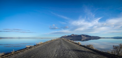 A long, empty road stretches towards mountains, reflecting in calm water under a clear blue sky with wispy clouds.