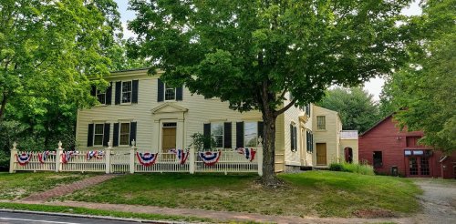 A historic yellow house with black shutters, surrounded by greenery and decorated with red, white, and blue bunting.