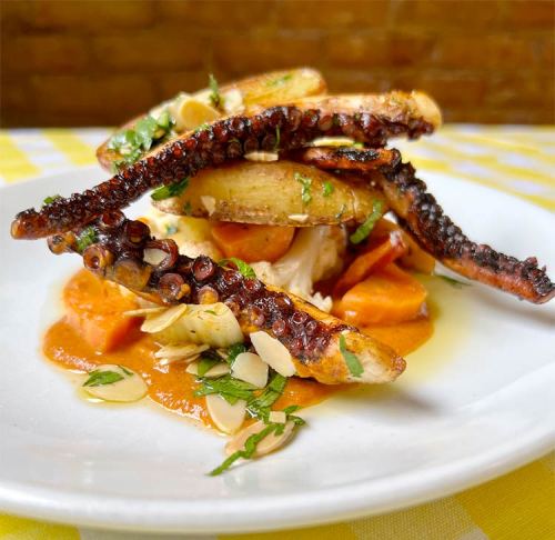 A plate of octopus tentacles atop roasted vegetables, garnished with herbs and sliced almonds on a vibrant sauce.
