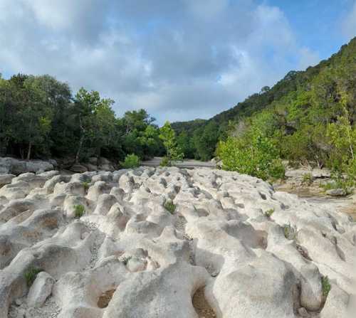 A rocky landscape with uneven terrain, surrounded by greenery and trees under a partly cloudy sky.